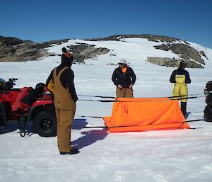 Orange rectangular tent roped between two red quad bikes. On white snow covered ground, leading to hill with snow and rock in distance with blue sky above. Three expeditioners staning around the bivvy bag.