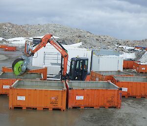 Five orange open topped 1/4 height shipping containers placed around an orange digger. In distance more containers, rocky hill and grey sky.