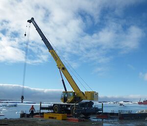 Front centre picture — large yellow crane with arm up into sky with rigging hanging down. In the distance in the bay is an orange ship. Blue skies with white cloud above.