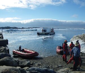 Whaft to left of picture leading to stoney boat ramp. Two red IRB are tied up at boat ramp. In the distance, large barge with white shipping container embarked is heading out into the bay. In front right of picture are four expeditioners in red and blue waterproof full length suits