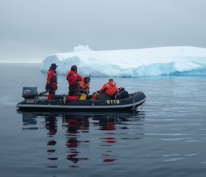 Grey still water to horizon, grey sky above. Centre picture a black IRB holding four expeditioners is floating past a small iceberg.