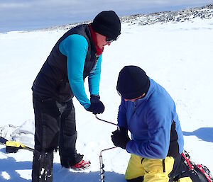 Two expeditioners drilling the sea-ice with hand held auger. Expeditioner on left is standing wearing black pants, vest, beanie and aqua jumper. Expeditioner on right is kneeling wearing yellow and black pants, blue jacket and black beanie. White ice to horizen. Blue sky above.