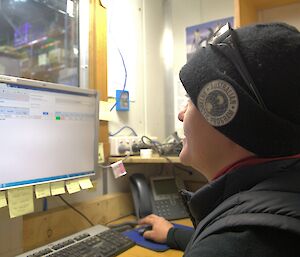 Close in shot, woman on right from shoulders up wearing black vest and beanie. Looking at computer screen on left and holding a mouse. In background through window is the warehouse.