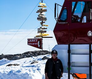 Front of large red and white bus filling right side of picture. In front, coming up to bumper bar is a woman wearing black. Background is snow covered hill with Casey direction/distances sign and blue sky.