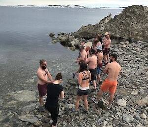 Group of about 12 swimmers on edge of water with rocky ground underneath, icebergs in distance