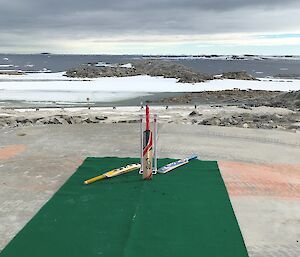 Cricket pitch consisting of green carpet roll, metal stumps and three cricket bats on cement helipad. In the distance is a bay with blue water and icebergs.