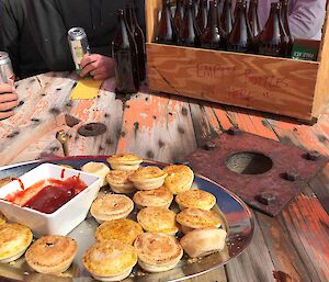 On top of large wooden barrel is a tray of party pies and bowl of tomato sauce. At back is a crate with brown beer bottles.