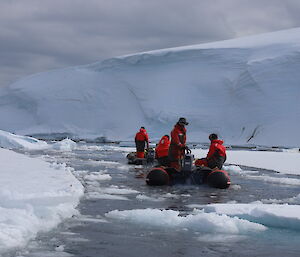 Two red boats with three people in each (wearing red and black mustang suits) moving through channel of water in sea ice. In the distance are large ice cliffs.