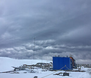 Bottom third is snow covered slope with blue square building on right and metal piping for site services leading to building. Top two thirds is sky with grey clouds and patch across centre picture is mammatus cloud which has a portion drooping down towards the ground like udders.