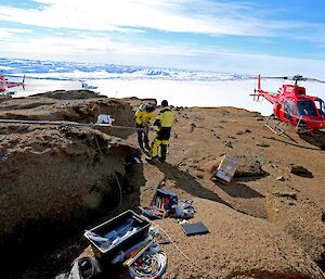 Rocky outcrop with two red helicopters landed to far left and right. In centre two men in yellow and black outdoor gear are working on a dropped metal mast with boxes of equipement open in front.