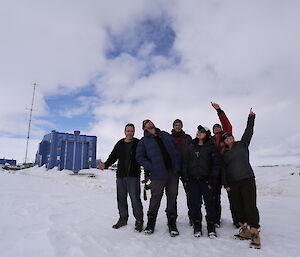Group of six people dressed in black pointing to the sky at front right of picture. White snow covered ground to horizen mid picture. Blue building on horizen at left of picture. Cloud covered sky above.