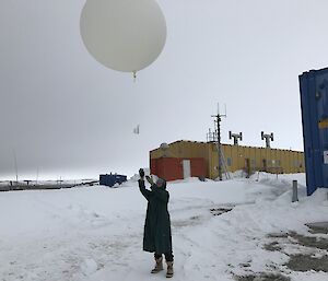 Centre picture person in lab coat holding a large round white balloon. On snow covered ground to horizen and grey sky above. To right of picture is edge of blue building and behind a yellow rectangular building