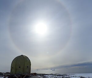 Foreground, dome shaped building in rusted yellow on rocky ground. Sky is light grey with a circular halo around the sun just to left of centre of picture.