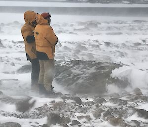 Full frame left of picture two men sanding in yellow puffer jackets and brown overpants. Rest of picture if snow covered rocky ground with blowing snow causing reduced visibility