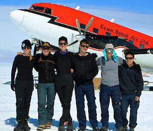 A group of six expeditioners standing with arms across each others shoulders, behind the front portion of a DC-3 (Basler) aircaft painted white underneath and red on top.
