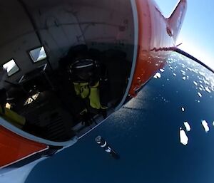 Fish lens image of the open door of an aeroplane in flight with man in yellow and black winter clothing at the opening. Underneath is the blue ocean with speckles of white icebergs and far right corner is blue sky