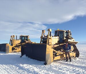 Two large yellow snow ploughs, one centre picture and one to back left on snow runway to horizen at mid picture. Blue sky with white clouds filling top left above. Man in brown overpants and blue and high vis jacket standing in front of first plough