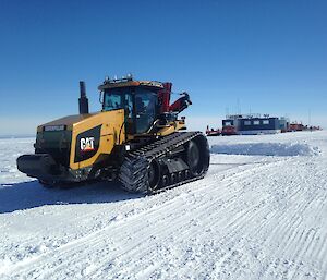 Large yellow tracked tractor pulling a plough behind, grades snow runway. In the distance is a blue building made from shipping container. Blue sky above.
