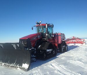 Large red tracktor with plough at front and draging red metal land plane behind. On flat snow runway with blue sky above.