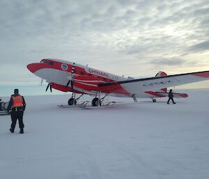 Snow runway, cloud covered sky, red and white DC3 basler aircraft mid picture, with person in black winter clothes and high vis vest at front left.