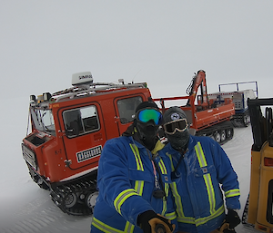 White background with red hagglunds. Two men standing in front with heavy blue jackts with high vis markings, balaclavas, beanies and goggles.
