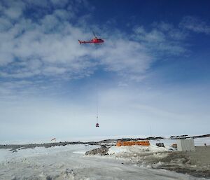 Blue sky with white clouds, foreground snow and rock covered. Collection of yellow fuel drums centre picture. Top centre a red helicopter carries below a long net with fuel drums inside.
