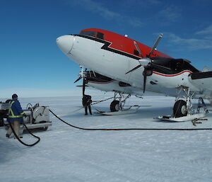 Top right of picture, red and white plane (forward of wings). On white snow runway. Blue sky above. Two men standing at fuel pump to left of picture with hose running towards the plane