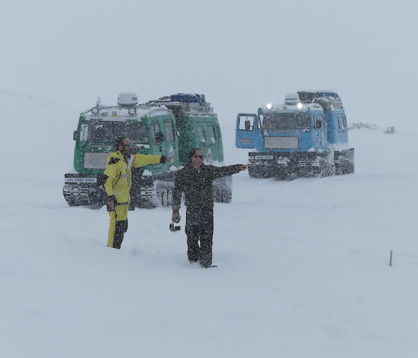 White out with green and blue Hägglunds vehicles seen through the snow. In front two men standing in the snow, both pointing to the right.
