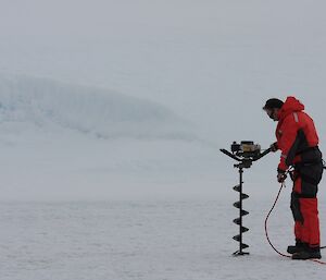 Ice drilling on flat ice covered lake, White lake surface to white ice cliffs, man in mustang suit with motorised large ice drill on right of picture