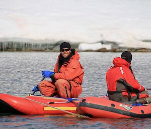 Two men in red blow up rafts connected together rowing on lake with ice covered edge in background