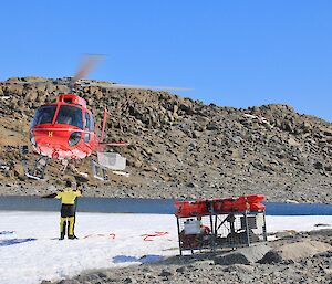 Helicopter hovering approx 15 feet above ground with man underneath directing sling loading operation. In foreground is a cage pallet prepared to be sling loaded