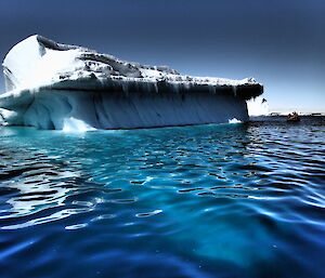 Aqua water with ripples in foreground to horizen. On horizen large tipped over iceberg with icicles along edge. Blue sky above