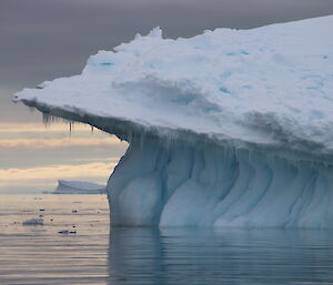 Sunset in distance, light tinged sky and sea. Large iceberg with mushroom shaped top to right of picture