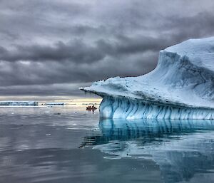 Right — large jade iceberg reflected onto water. Mid picture small RHIB in the distance. Grey sky above.