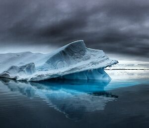 Ominous grey clouds, grey mirrored water. Mid picture mis-shapen iceberg with intense jade colours reflected into water