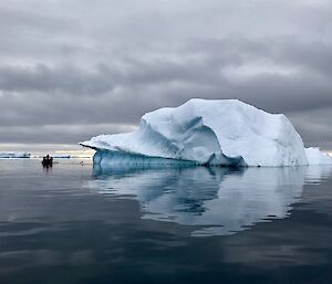 Horizen mid picture, grey sea and grey sky. Iceberg mid picture with reflection in mirrored water. Two small boats mid left