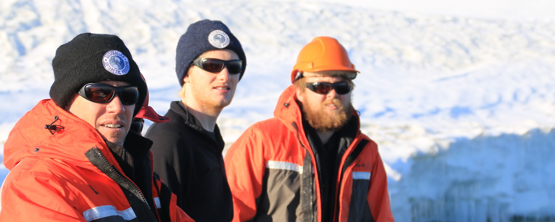 Three expeditioners in mustang suits, glasses and beanies in foreground, behind snow covered slope with buildings in distance