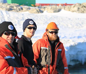 Three expeditioners in mustang suits, glasses and beanies in foreground, behind snow covered slope with buildings in distance