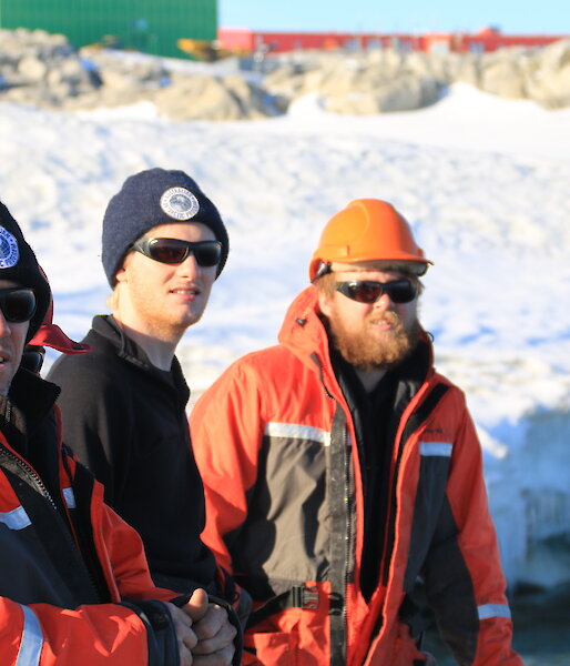 Three expeditioners in mustang suits, glasses and beanies in foreground, behind snow covered slope with buildings in distance
