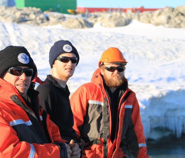 Three expeditioners in mustang suits, glasses and beanies in foreground, behind snow covered slope with buildings in distance