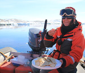 Lady sitting at end of IRB in mustang suit and with goggles on head, holding a platter of cheese and biscuits with water and rocky slope behind