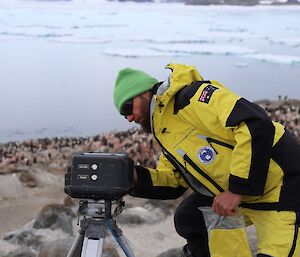 Expeditioner in black and yellow AAD cold weather gear and green beanie looking at large digital camera. In background penguin rookery and sea with sea-ice