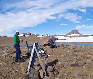Seismometer in foreground on rocky ground. In middle distance two expeditioners, one collecting rocks. In background a blue melt lake, rocky mountains and blue sky