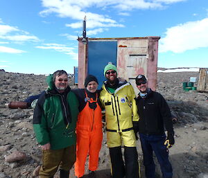 Four expeditioners standing linking arms. Behind an old run down field hut on rocky gound