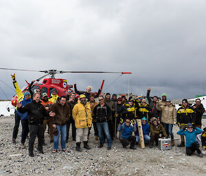 A large group of expeditioners standing in front of a red helicopter. On rocky surface with grey sky above
