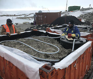 A quarter height orange container filled with soil and a water pipe coiled across the surface. Two expeditioners at rear of container looking over the edge