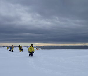 Landscape shot of white snow covered ground to grey sea and grey stormy clouds above. A group of expeditioners spread out walking through the snow