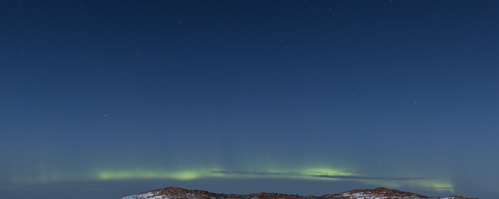 Bottom third of photo is rocky and snow covered hill, just above is a pale yellow / green aurora and then rest of photo is a blue black night sky with stars scattered across