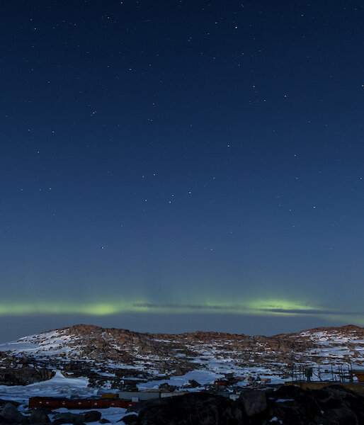 Bottom third of photo is rocky and snow covered hill, just above is a pale yellow / green aurora and then rest of photo is a blue black night sky with stars scattered across