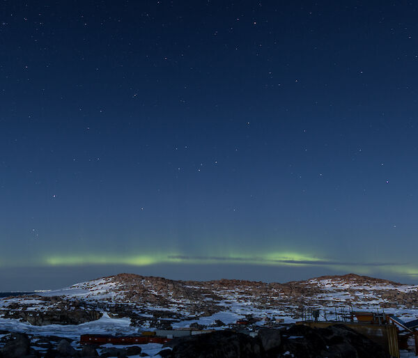 Bottom third of photo is rocky and snow covered hill, just above is a pale yellow / green aurora and then rest of photo is a blue black night sky with stars scattered across